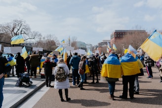 A group of people gathered in an outdoor space, many draped in yellow and blue flags, resembling the Ukrainian flag. They are holding signs and flags, participating in what appears to be a protest or demonstration. The background shows leafless trees and buildings, indicating a possible urban location in cooler weather.