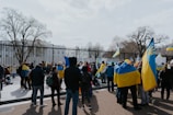 A group of people gathered outside, holding and waving blue and yellow flags in front of a black metal fence. Leafless trees are visible in the background, suggesting a cold season.