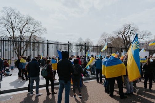 A group of people gathered outside, holding and waving blue and yellow flags in front of a black metal fence. Leafless trees are visible in the background, suggesting a cold season.