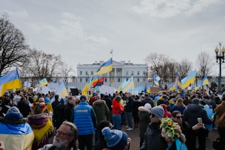A large group of people gathered in front of a notable white building, holding Ukrainian flags and signs. Many are wearing winter clothing, and the scene is surrounded by leafless trees. The mood suggests a peaceful protest or demonstration.