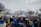 A large group of people gathered in front of a notable white building, holding Ukrainian flags and signs. Many are wearing winter clothing, and the scene is surrounded by leafless trees. The mood suggests a peaceful protest or demonstration.