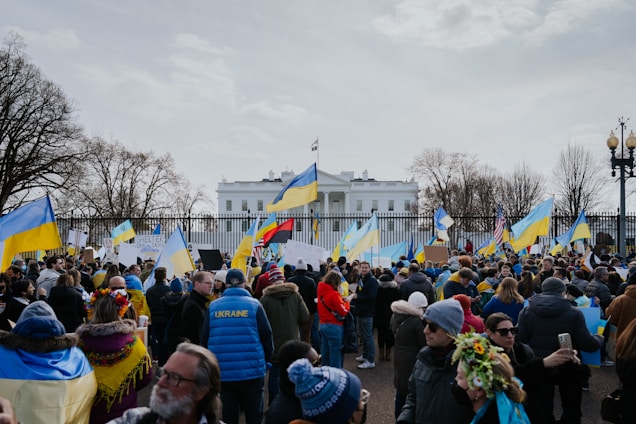 A large group of people gathered in front of a notable white building, holding Ukrainian flags and signs. Many are wearing winter clothing, and the scene is surrounded by leafless trees. The mood suggests a peaceful protest or demonstration.