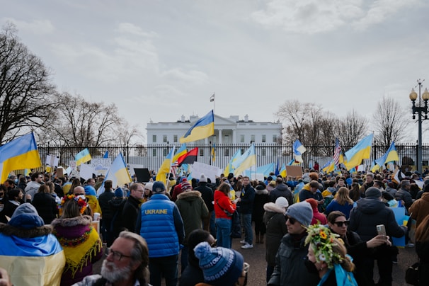 A large group of people gathered in front of a notable white building, holding Ukrainian flags and signs. Many are wearing winter clothing, and the scene is surrounded by leafless trees. The mood suggests a peaceful protest or demonstration.