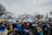A large group of people gathered in front of a notable white building, holding Ukrainian flags and signs. Many are wearing winter clothing, and the scene is surrounded by leafless trees. The mood suggests a peaceful protest or demonstration.
