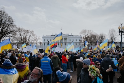 A large group of people gathered in front of a notable white building, holding Ukrainian flags and signs. Many are wearing winter clothing, and the scene is surrounded by leafless trees. The mood suggests a peaceful protest or demonstration.
