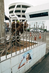A modern circular multi-story parking structure is visible behind a metal fence with pointed tops. In front of the fence, there is a poster of a person drinking water on a tiled wall. The environment appears urban with underdeveloped vegetation and traffic cones visible nearby.