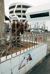 A modern circular multi-story parking structure is visible behind a metal fence with pointed tops. In front of the fence, there is a poster of a person drinking water on a tiled wall. The environment appears urban with underdeveloped vegetation and traffic cones visible nearby.