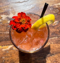 A refreshing pink beverage in a glass garnished with a vibrant red flower and a lemon slice. It has two black straws and is placed on a rustic wooden surface with sunlight casting distinctive shadows.