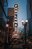 A city street scene features a large, illuminated vertical sign for 'PORTLAND' in vertical letters. The sign is adorned with bright lights and stands out against the early evening sky. Below it, a smaller marquee displays event details for a literary arts presentation. The street is populated with pedestrians and surrounded by modern buildings, lampposts, and some trees adorned with lights.