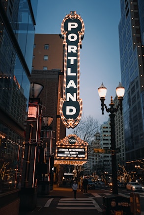 A city street scene features a large, illuminated vertical sign for 'PORTLAND' in vertical letters. The sign is adorned with bright lights and stands out against the early evening sky. Below it, a smaller marquee displays event details for a literary arts presentation. The street is populated with pedestrians and surrounded by modern buildings, lampposts, and some trees adorned with lights.
