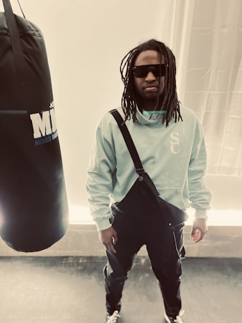 A person with long hair wearing sunglasses and casual clothing stands in a room with a large punching bag labeled 'MMA' to the side. The person is dressed in a light-colored hoodie with a strap across the chest and dark pants.