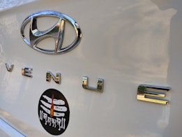 Close-up of a car trunk with a shiny silver Hyundai emblem and the word 'VENUE' in chrome letters. Below these, a black oval sticker features a white Shiva linga with a flame design and Devanagari script.