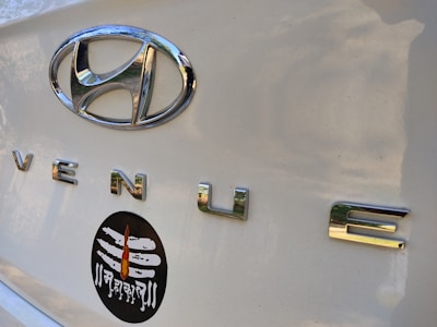 Close-up of a car trunk with a shiny silver Hyundai emblem and the word 'VENUE' in chrome letters. Below these, a black oval sticker features a white Shiva linga with a flame design and Devanagari script.