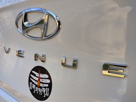 Close-up of a car trunk with a shiny silver Hyundai emblem and the word 'VENUE' in chrome letters. Below these, a black oval sticker features a white Shiva linga with a flame design and Devanagari script.