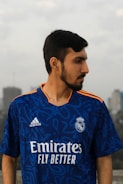 A person with a beard is wearing a blue sports jersey with the Real Madrid logo and sponsor branding. They are outdoors, with a blurred urban background and cloudy sky.