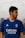 A person with a beard is wearing a blue sports jersey with the Real Madrid logo and sponsor branding. They are outdoors, with a blurred urban background and cloudy sky.