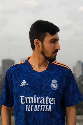 A person with a beard is wearing a blue sports jersey with the Real Madrid logo and sponsor branding. They are outdoors, with a blurred urban background and cloudy sky.