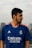 A person with a beard is wearing a blue sports jersey with the Real Madrid logo and sponsor branding. They are outdoors, with a blurred urban background and cloudy sky.