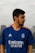 A person with a beard is wearing a blue sports jersey with the Real Madrid logo and sponsor branding. They are outdoors, with a blurred urban background and cloudy sky.