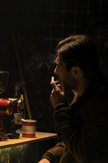 A person is sitting in a dimly lit room, holding a cigarette with smoke rising. A coffee grinder and a red and white coffee tin are visible on the table. The atmosphere is moody and contemplative.