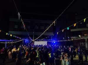 A large group of people gathers outdoors at night in an open area, likely for a celebration or party. String lights and colorful flags are hung overhead, casting a festive glow. The crowd appears to be dancing or mingling in front of a building with blue lighting enhancing the party atmosphere.