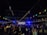 A large group of people gathers outdoors at night in an open area, likely for a celebration or party. String lights and colorful flags are hung overhead, casting a festive glow. The crowd appears to be dancing or mingling in front of a building with blue lighting enhancing the party atmosphere.