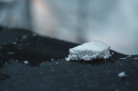 A small piece of white foam lies on a dark, textured surface. The foam appears jagged and uneven, with small spherical protrusions typical of styrofoam. The background is softly blurred, dominated by cool colors, lending an abstract and minimalistic feel.