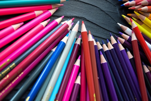 A collection of brightly colored pencils arranged in a circular formation on a dark textured surface. The pencils display a variety of vibrant shades, including reds, pinks, blues, purples, and yellows.
