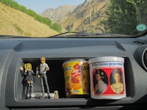 A car dashboard compartment contains several items: two anime figurines, a large tin labeled 'Wilhelmina pepermunt' with historical portrait images, and a box of 'BNIZ Minis' snacks. The dashboard is gray and the vehicle is parked with a view of mountainous terrain and green foliage outside the window.