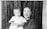 A woman and a young child pose together in front of a textured backdrop. The woman has a calm expression and is wearing a patterned dress, while the child, dressed in a white garment, looks towards the camera with a subtle, slightly blurred expression.