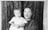 A woman and a young child pose together in front of a textured backdrop. The woman has a calm expression and is wearing a patterned dress, while the child, dressed in a white garment, looks towards the camera with a subtle, slightly blurred expression.