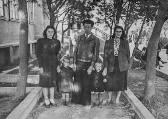 A black-and-white photograph featuring five people, possibly a family, standing outdoors. The group consists of three adults and two children. They are positioned on a path surrounded by trees and greenery, with a building visible in the background.