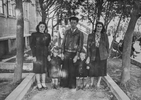 A black-and-white photograph featuring five people, possibly a family, standing outdoors. The group consists of three adults and two children. They are positioned on a path surrounded by trees and greenery, with a building visible in the background.