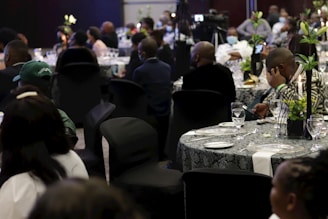 A formal event taking place in a dimly lit venue. Multiple round tables are set with tableware and decorated with floral centerpieces. Attendees are seated in black chair covers, wearing formal attire, some engaged in conversation while others appear to be listening or using their phones.