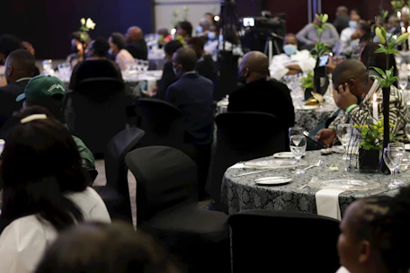 A formal event taking place in a dimly lit venue. Multiple round tables are set with tableware and decorated with floral centerpieces. Attendees are seated in black chair covers, wearing formal attire, some engaged in conversation while others appear to be listening or using their phones.