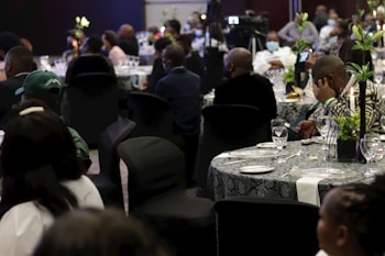 A formal event taking place in a dimly lit venue. Multiple round tables are set with tableware and decorated with floral centerpieces. Attendees are seated in black chair covers, wearing formal attire, some engaged in conversation while others appear to be listening or using their phones.
