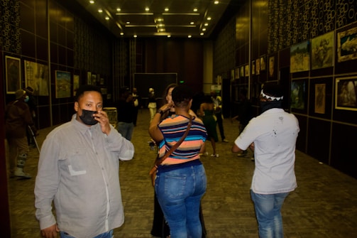 An indoor art gallery with several people walking and standing around. The walls are adorned with various framed artworks. The lighting is subdued with spotlights focusing on the paintings. Some individuals are wearing face masks.
