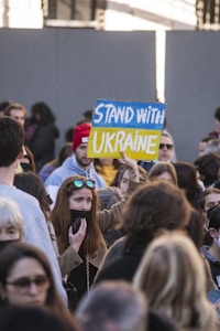 A large group of people gathered together, with one individual prominently holding up a sign that reads 'Stand with Ukraine'. The sign is painted in blue and yellow, reflecting the colors of the Ukrainian flag. Various expressions are seen on the faces of the crowd, with some people wearing coats and others having accessories like sunglasses and hats.