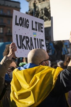 A crowd holds a sign reading 'Long Live Ukraine' with small red hearts and a heart in blue and yellow colors. A person, wearing a blue and yellow garment, is holding the sign high. The background shows a blurred statue and a building.
