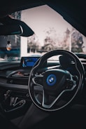 The image captures the interior of a car, focusing on the dashboard and steering wheel with a visible BMW logo. The dashboard features a digital display and various control buttons. Ambient lighting provides a sleek, modern look. The view through the windshield shows a blurry outdoor setting with some vegetation visible.