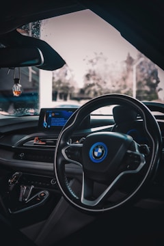 The image captures the interior of a car, focusing on the dashboard and steering wheel with a visible BMW logo. The dashboard features a digital display and various control buttons. Ambient lighting provides a sleek, modern look. The view through the windshield shows a blurry outdoor setting with some vegetation visible.