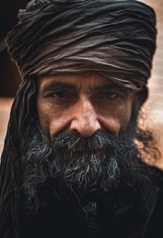 A man with a long, dark beard and a turban on his head, gazing directly into the camera. The wrinkles on his face and his intense eyes convey a sense of wisdom and experience. The lighting is subtle, highlighting the texture of his skin and beard.