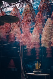 Rows of red spiral incense coils hang from the ceiling in an indoor space, creating a mesmerizing pattern. Below, a pot with burning incense sticks emits smoke that drifts upwards. The lighting is dim and atmospheric, giving the scene a mystical feel.