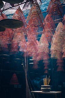 Rows of red spiral incense coils hang from the ceiling in an indoor space, creating a mesmerizing pattern. Below, a pot with burning incense sticks emits smoke that drifts upwards. The lighting is dim and atmospheric, giving the scene a mystical feel.