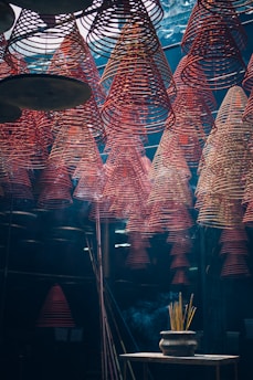 Rows of red spiral incense coils hang from the ceiling in an indoor space, creating a mesmerizing pattern. Below, a pot with burning incense sticks emits smoke that drifts upwards. The lighting is dim and atmospheric, giving the scene a mystical feel.