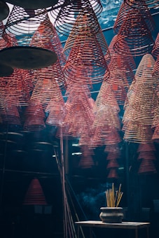Rows of red spiral incense coils hang from the ceiling in an indoor space, creating a mesmerizing pattern. Below, a pot with burning incense sticks emits smoke that drifts upwards. The lighting is dim and atmospheric, giving the scene a mystical feel.