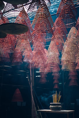Rows of red spiral incense coils hang from the ceiling in an indoor space, creating a mesmerizing pattern. Below, a pot with burning incense sticks emits smoke that drifts upwards. The lighting is dim and atmospheric, giving the scene a mystical feel.