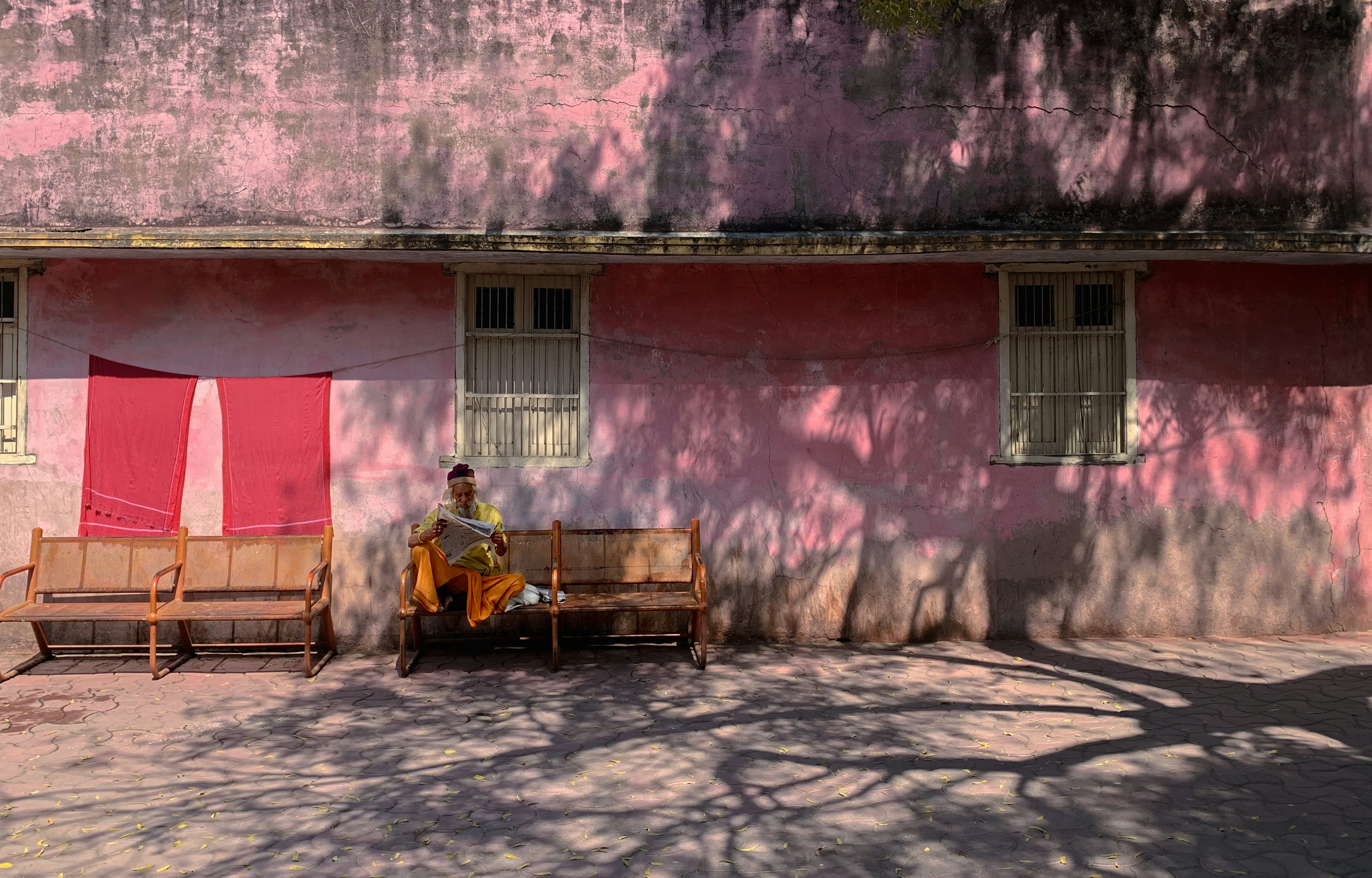 An old man reading newspaper sitting on a bench in front of a pink wall