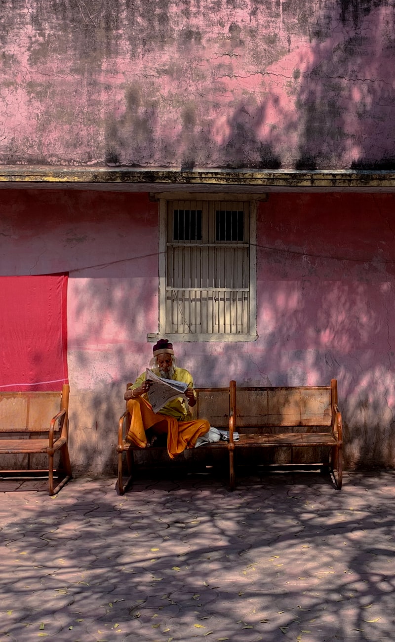 An old man reading newspaper sitting on a bench in front of a pink wall