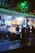 A group of people standing in front of an illuminated amusement arcade, engaging with various gaming machines. The environment is bustling with activity, and colorful lights create a vibrant atmosphere. The ceiling is decorated with large, playful images of dice and other game-related graphics, enhancing the lively mood.